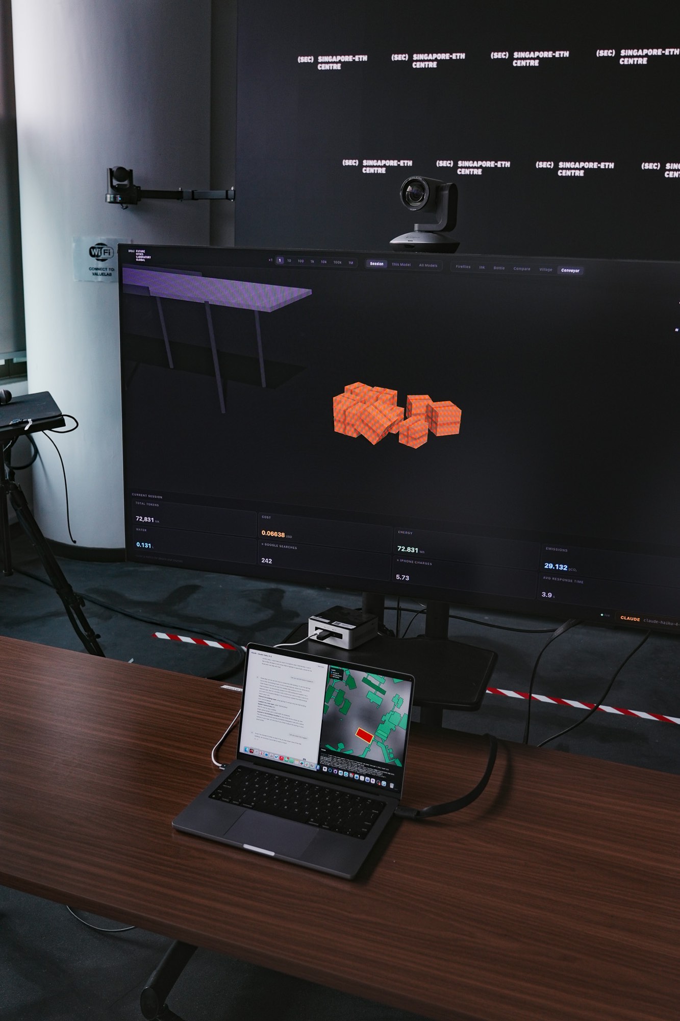 Close-up of the dual-screen exhibit setup showing the 3D geospatial visualization of building models on the main display with connected laptop showing the chatbot interface and map controls.